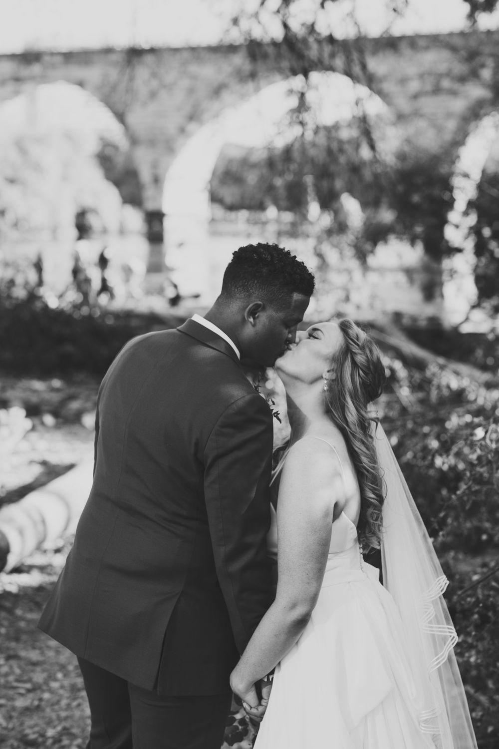 Black and white photo of bride and groom kissing near a stone arch bridge.