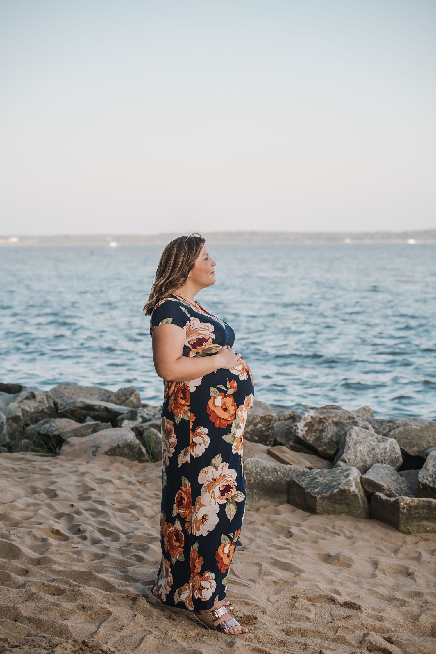 Person in floral dress sitting on coastal rocks at sunset overlooking the ocean.