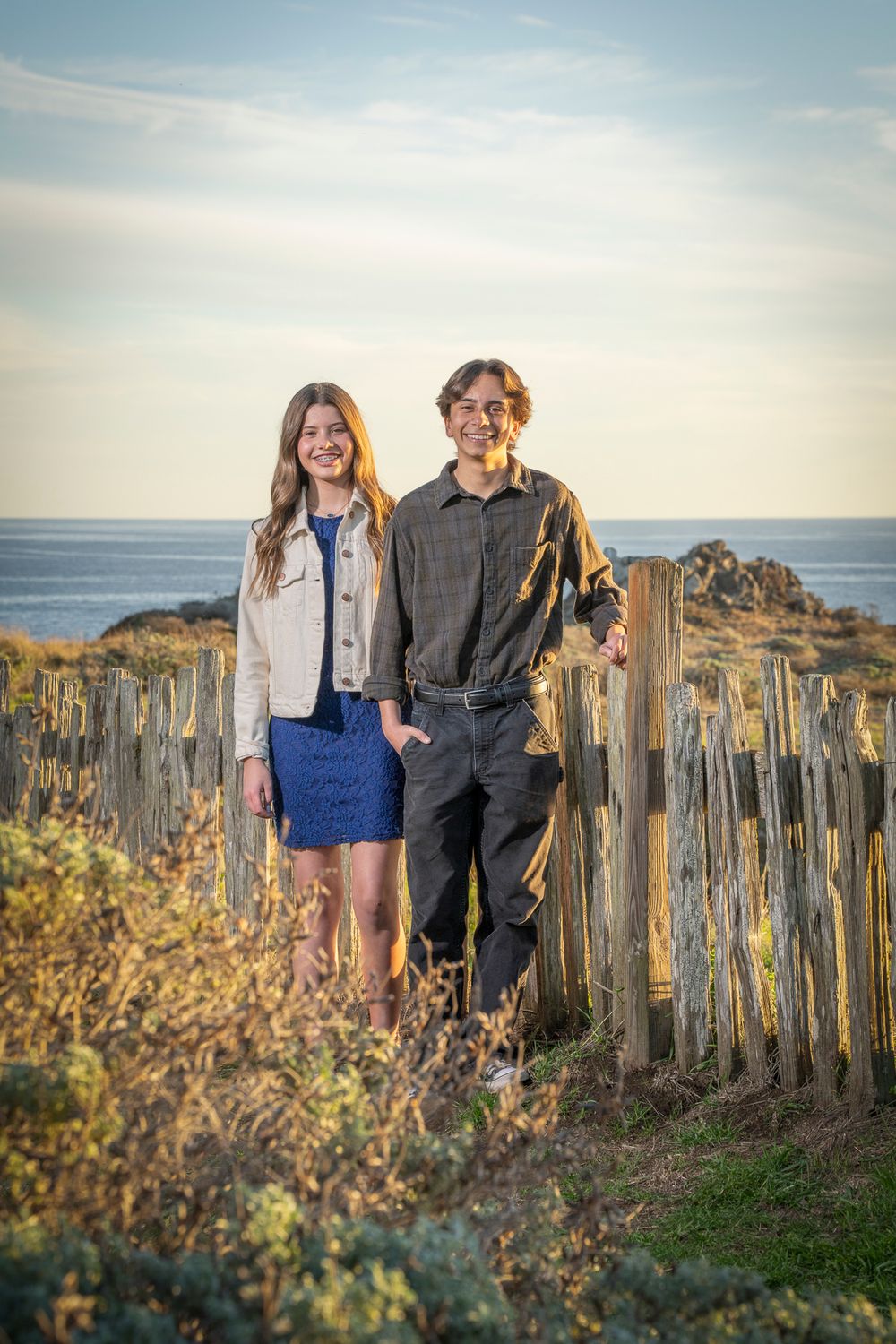 Couple standing by weathered fence posts against coastal landscape at sunset. At The Sea Ranch Lodge in Northern California.