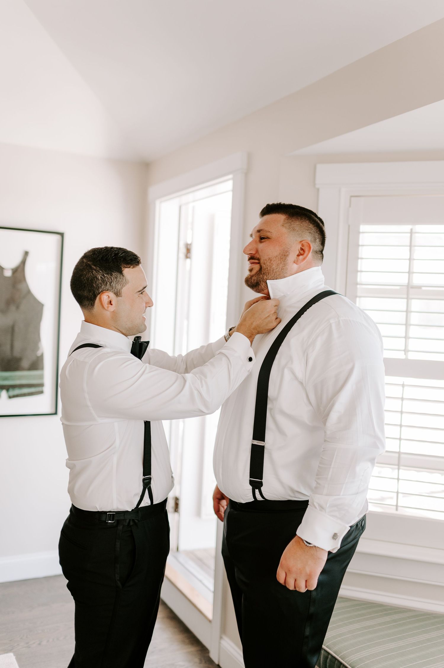 Two groomsmen in white shirts and suspenders help each other adjust their bow ties in a bright room.