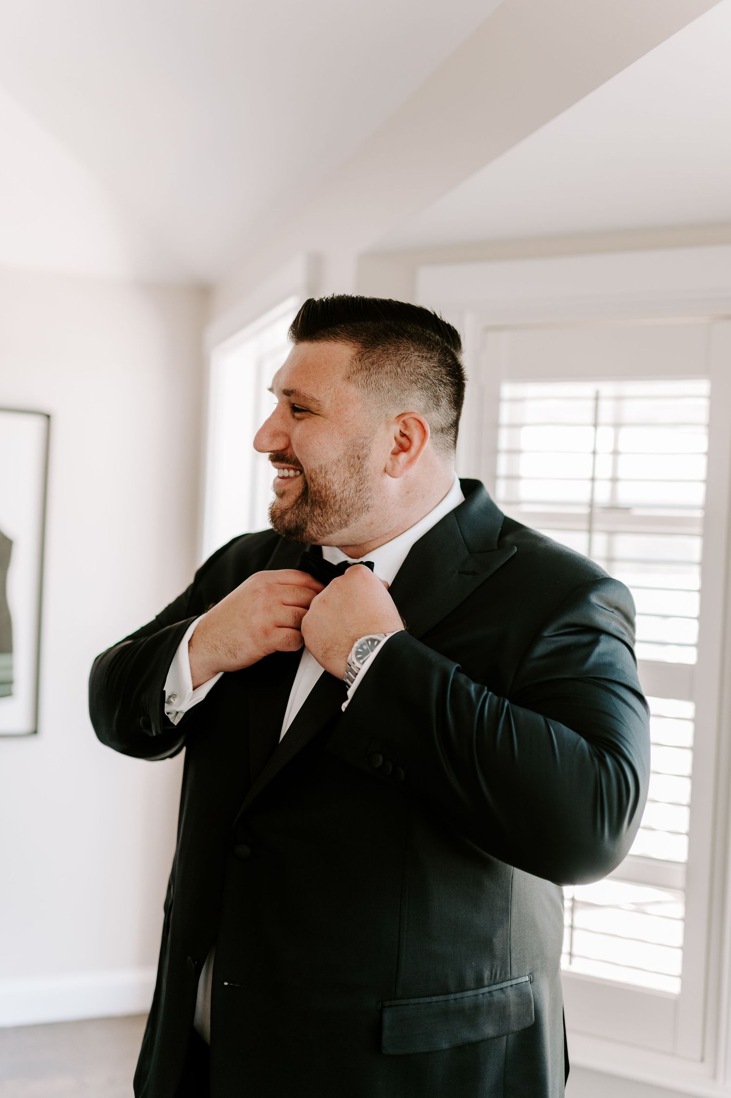 A groom in a black tuxedo adjusts his bowtie while smiling in a bright room.