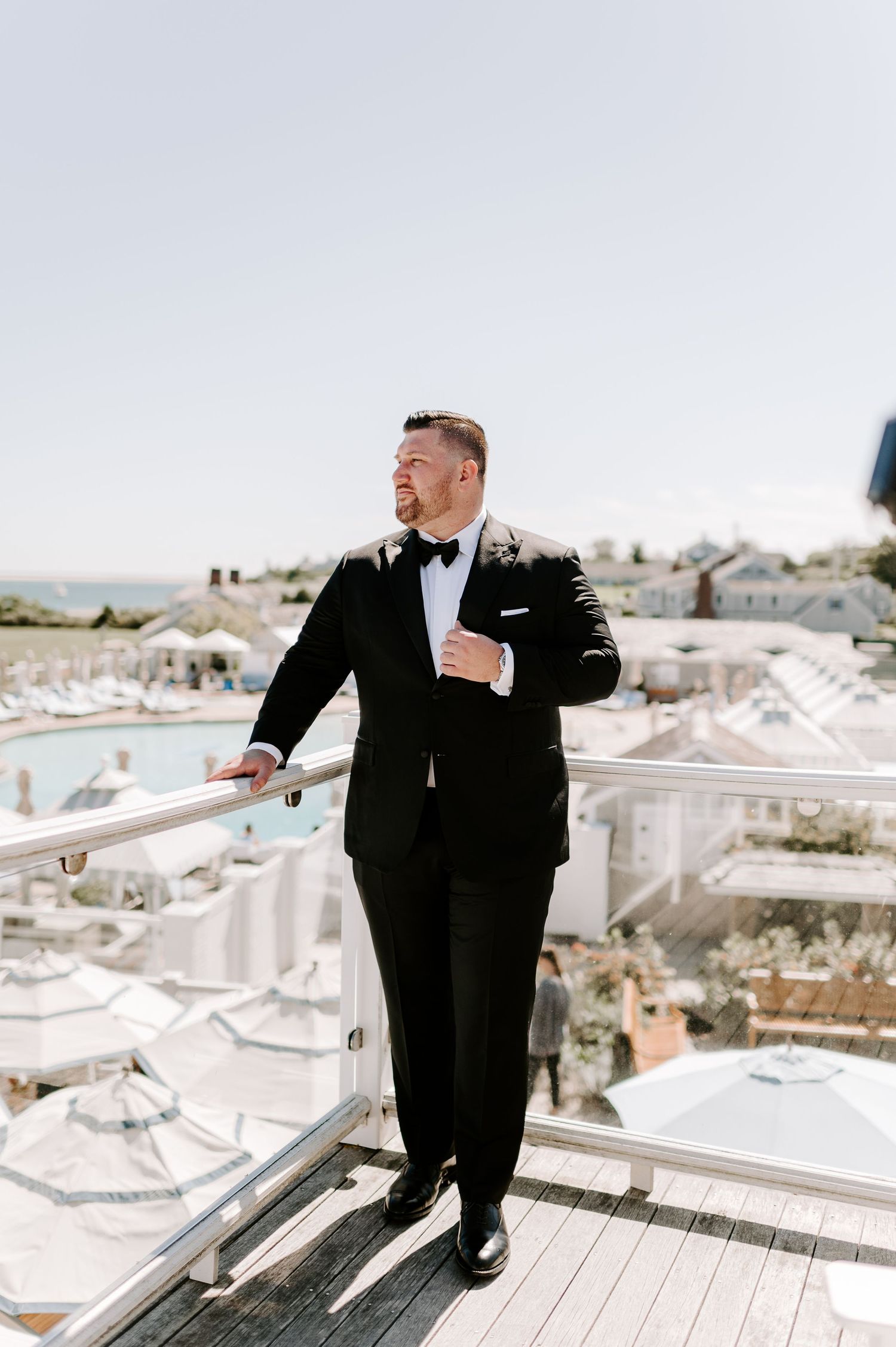 A groom in classic black tuxedo stands on balcony overlooking white coastal buildings.