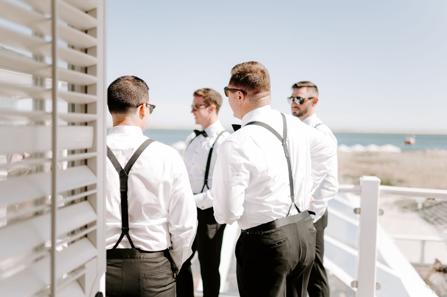 Groomsmen in white shirts and suspenders pose together on a sunny beach.