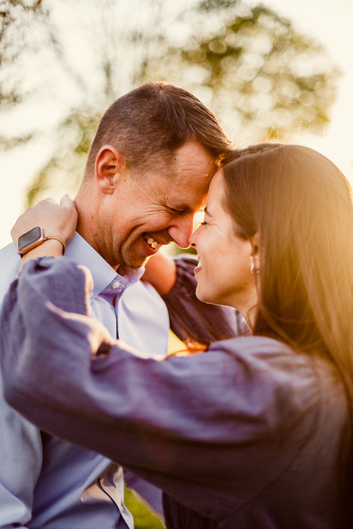 a couple stands forehead to forehead with golden sunset behind them, their eyes are closed and they are smiling happily