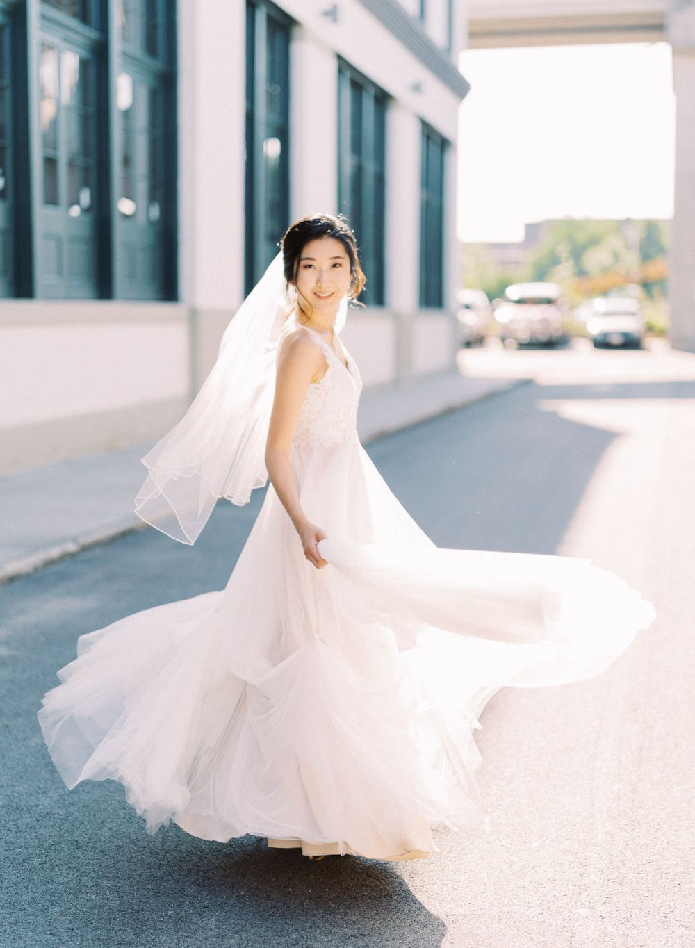 A bride in a flowing white gown twirls gracefully outside a modern building.