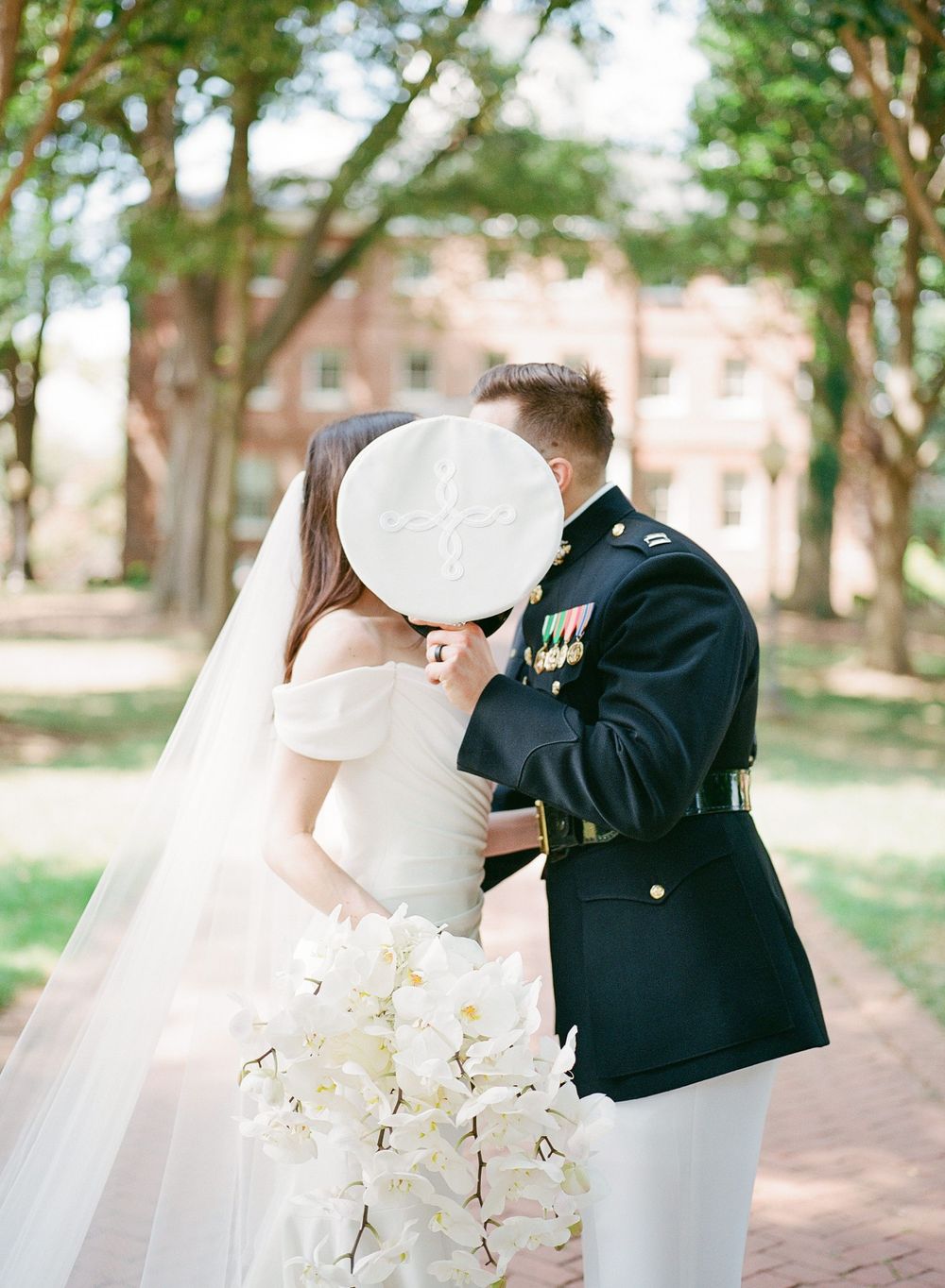 Wedding couple in white dress and military uniform embracing outdoors near a brick building.