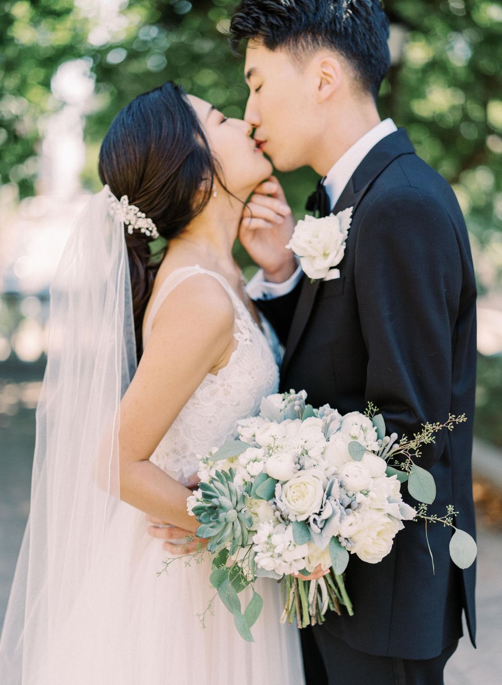 A tender wedding kiss captured outdoors with a pastel bouquet of roses and greenery.