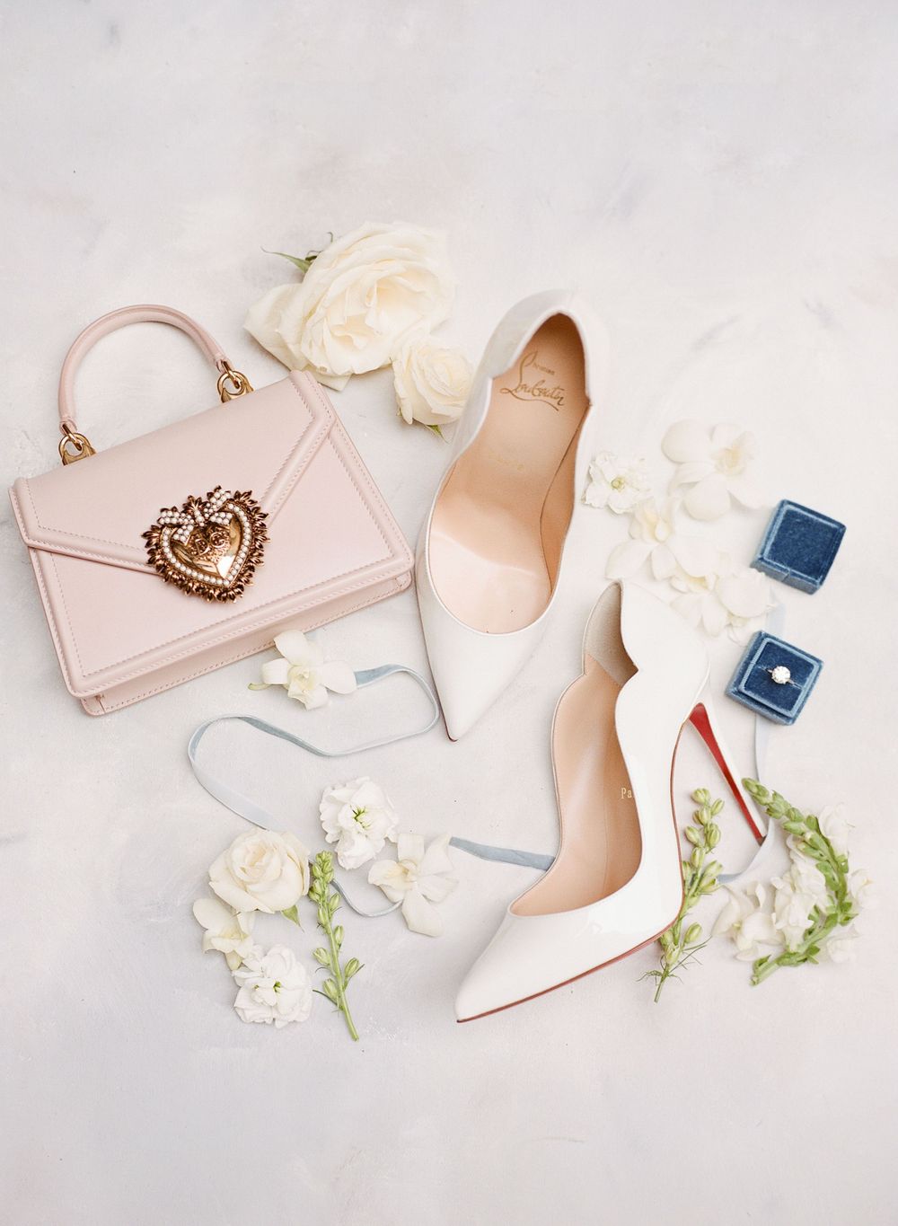 Bridal accessories flat lay with pink handbag, white heels, flowers and blue ring boxes.