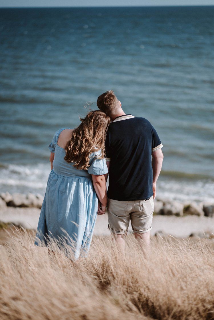 A couple stands together at the beach's edge, viewing the ocean through tall grass on a serene day.