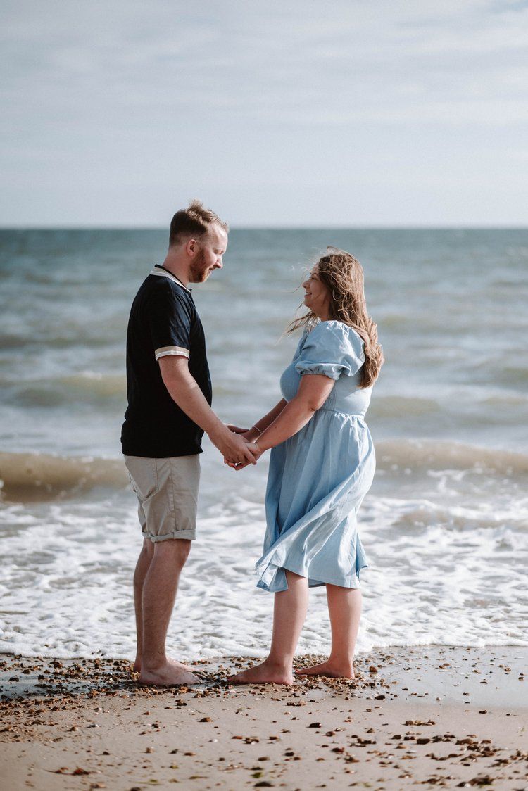 A couple stands holding hands on a beach with waves crashing behind them in a romantic seaside moment.