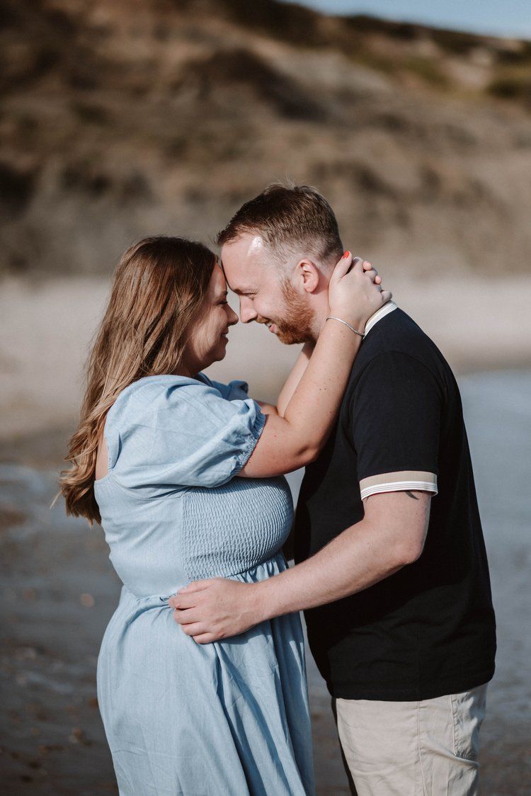 A romantic couple embraces on a beach at sunset, with the woman in a blue dress and man in dark shirt and khakis.