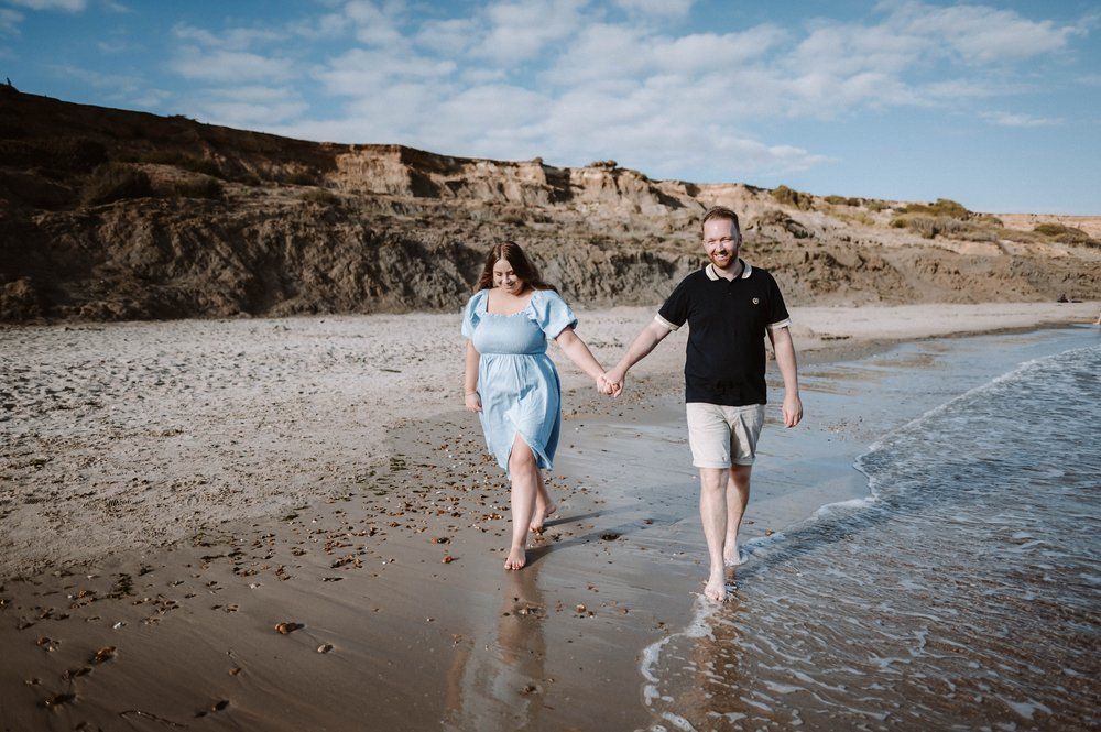 Two people holding hands while walking along the shoreline of a scenic beach with rugged cliffs in the background.