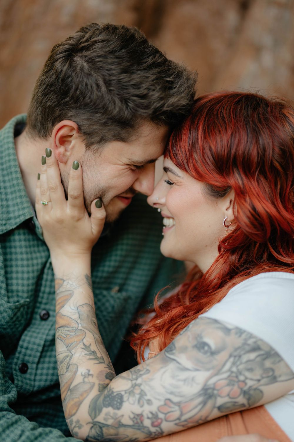 A close romantic moment between a couple with tattoos and red hair sharing laughter against a blurred brown background.