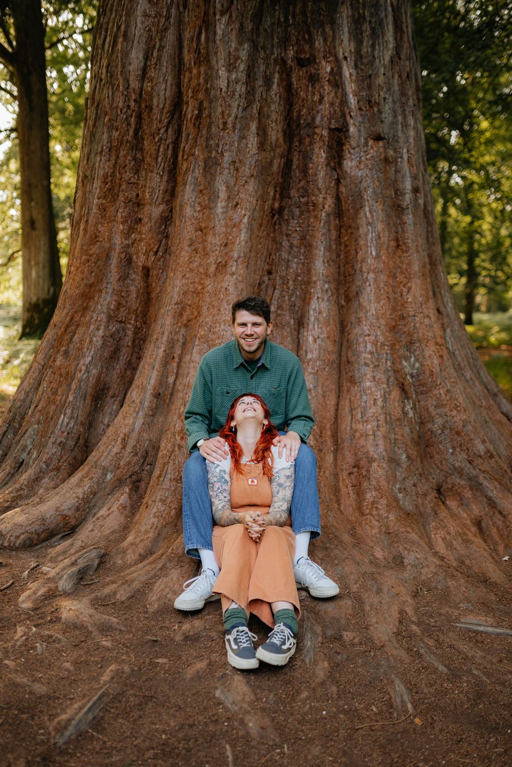 A couple sits together against a massive redwood tree trunk in a forest setting.