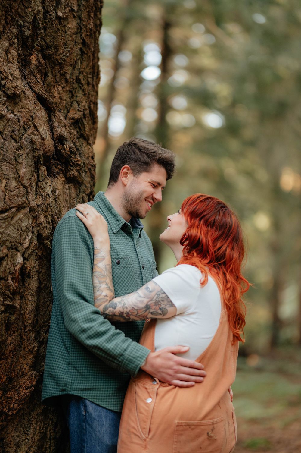 A couple shares a tender moment together while standing against a large tree trunk in a forest setting.