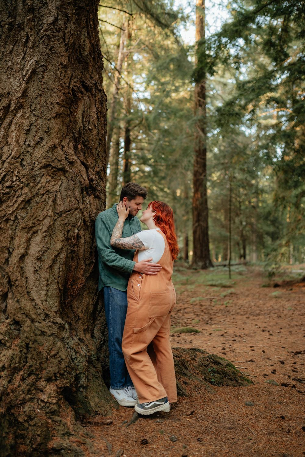 A romantic couple embraces against a large tree trunk in a peaceful forest setting with soft natural lighting.
