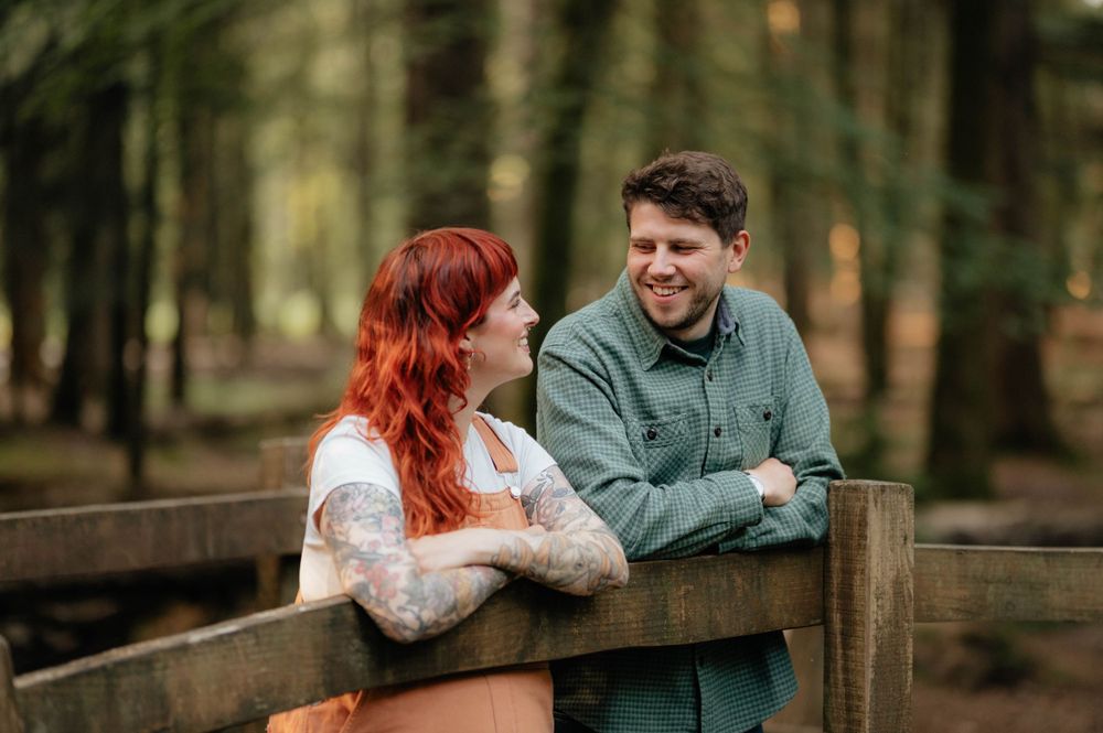 Couple leans on wooden bridge railing in forest during romantic outdoor engagement photoshoot.