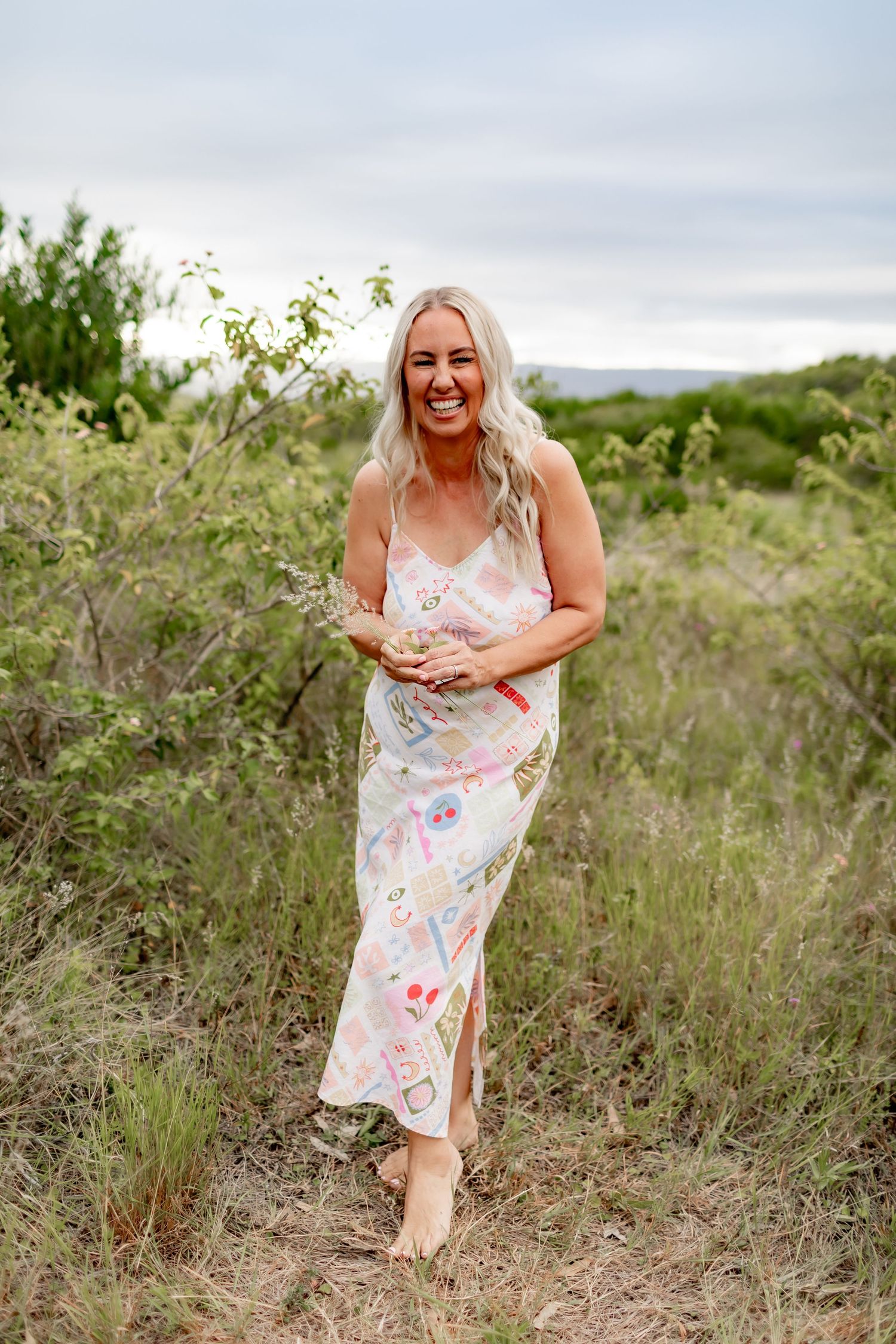 Woman in floral summer dress poses joyfully in a natural brushland setting under cloudy skies.