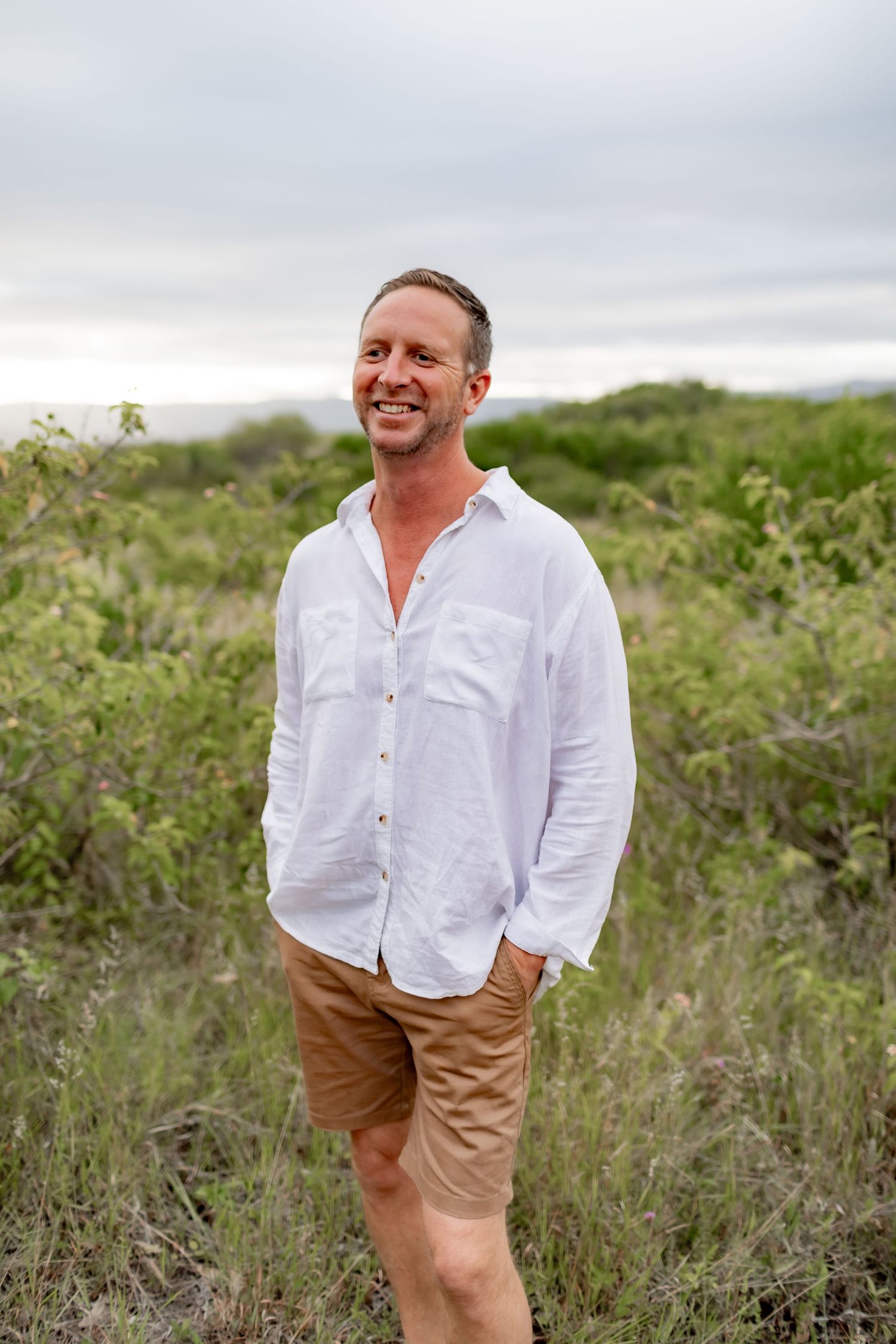 Man in white shirt and khaki shorts poses casually in brushland under cloudy skies.