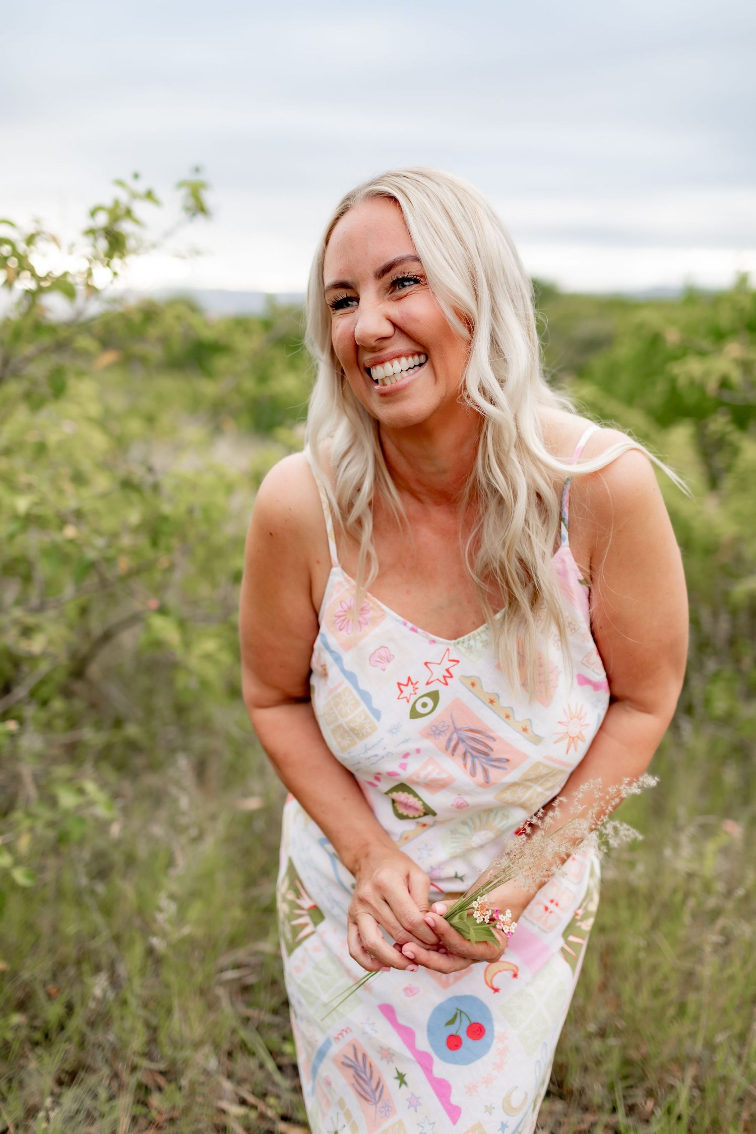 Woman wearing a floral dress laughs candidly while standing in wild brush under an overcast sky.