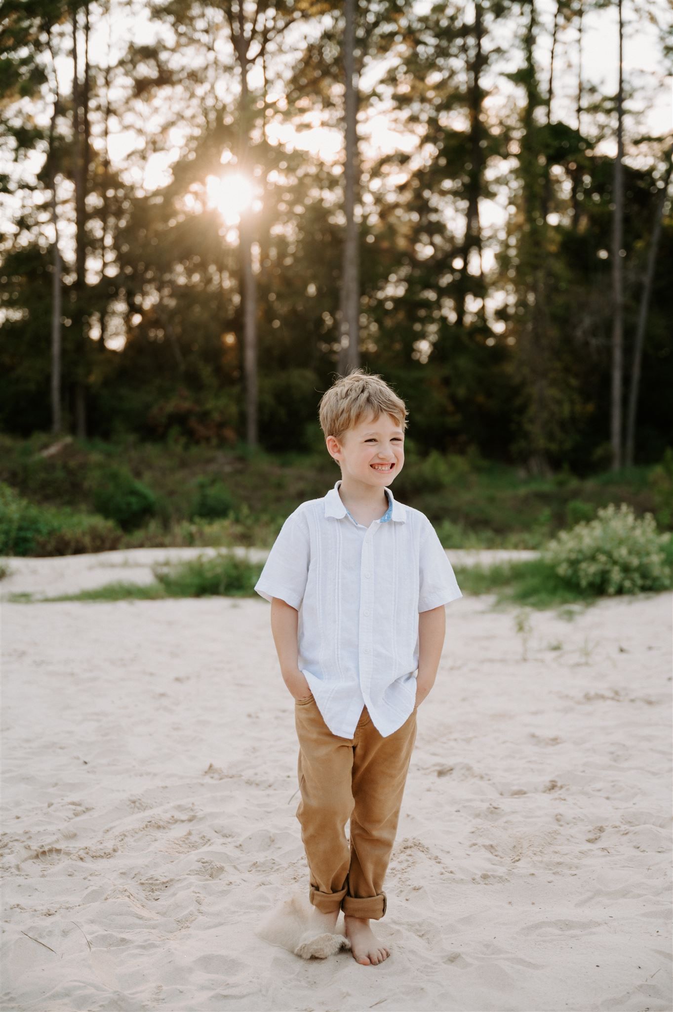Young boy in white shirt and khaki pants stands barefoot on sandy beach path.