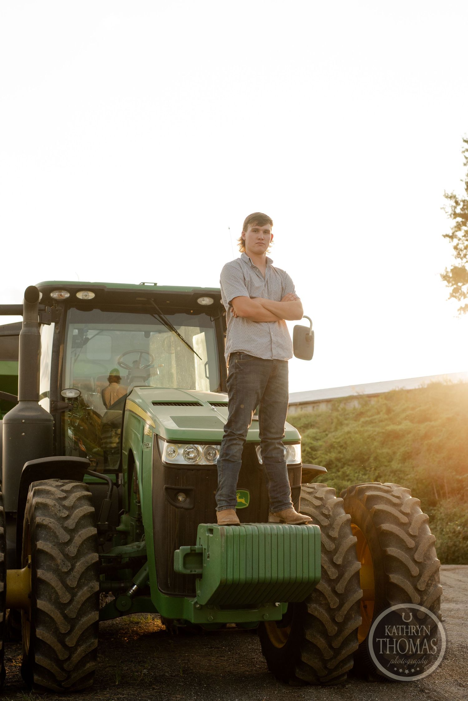 Person standing on green tractor against sunset sky in rural setting.