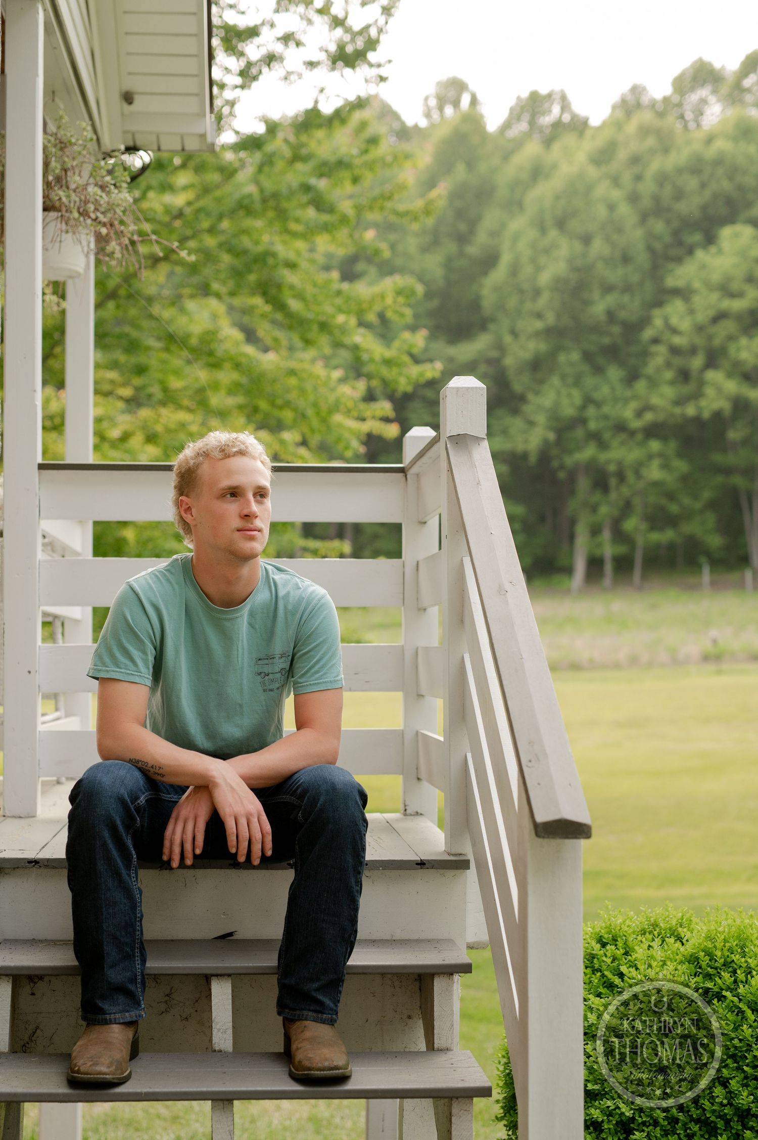Sitting on white porch steps wearing casual attire with wooded backdrop.