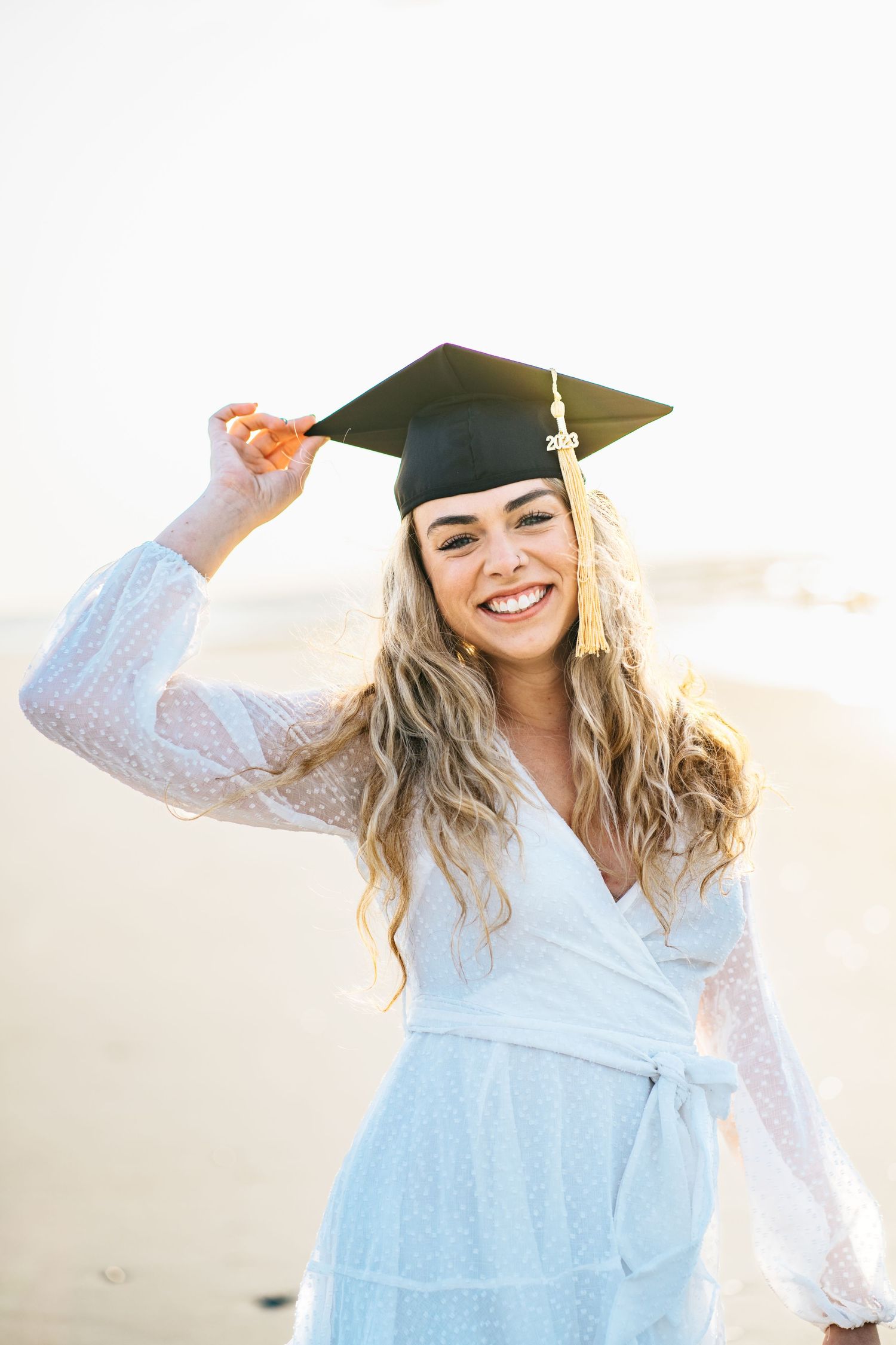 A graduating senior in a white dress holds her black cap with gold tassel smiling at the camera on the beach