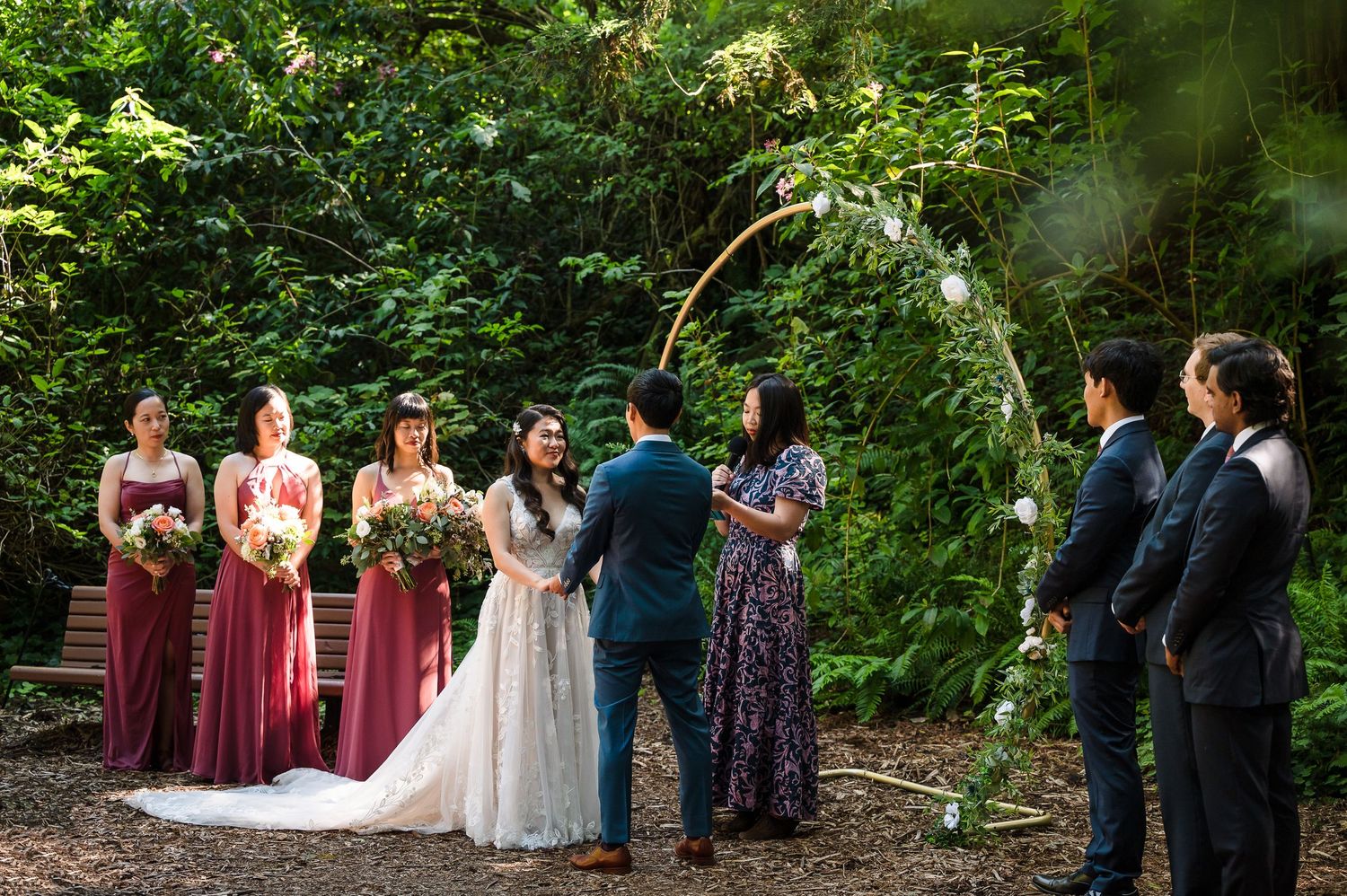 Outdoor wedding ceremony with burgundy-dressed attendants in a lush garden setting.