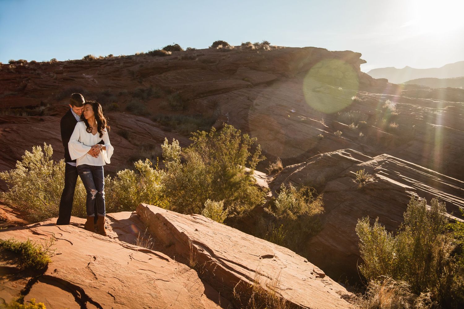 Couple embraces on red rock overlook surrounded by desert shrubs with sun flare and canyon views.