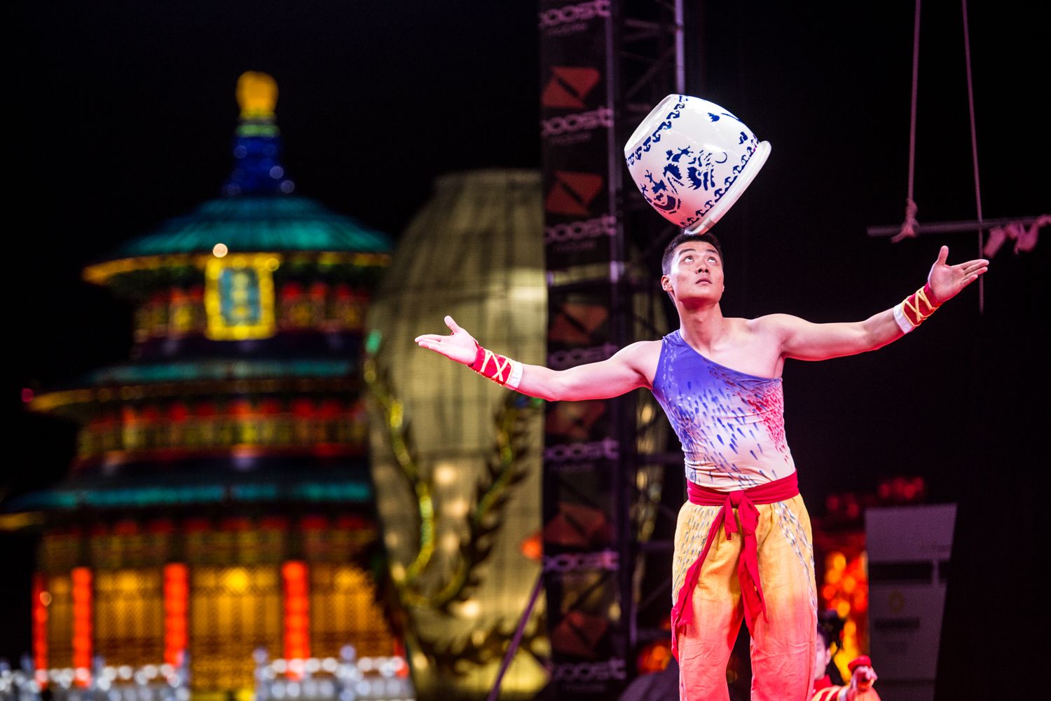 A performer balances a large dice cube while standing on stage at a circus show with colorful pagoda backdrop.