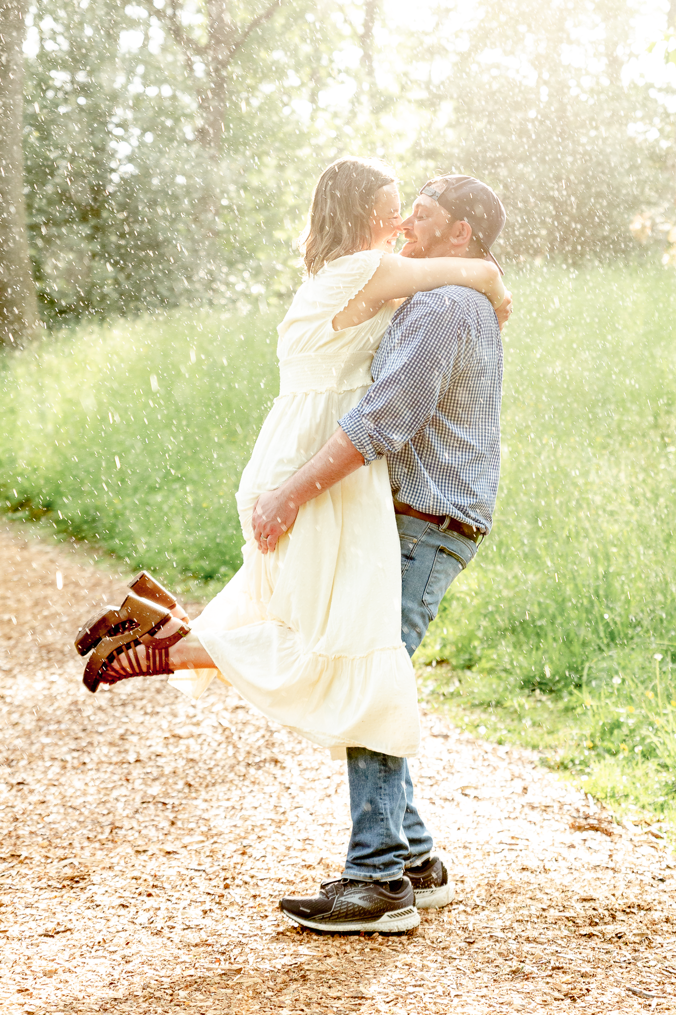 man in blue shirt holding woman in white dress kissing and spinning in the rain during golden hour sunlight
