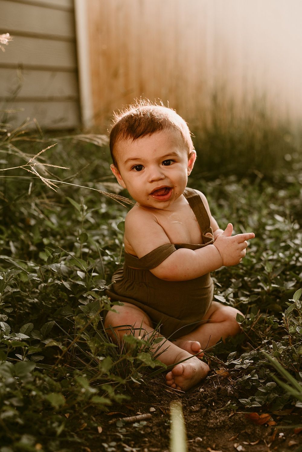 Baby in brown overalls sits in grass during golden hour backlit photoshoot.