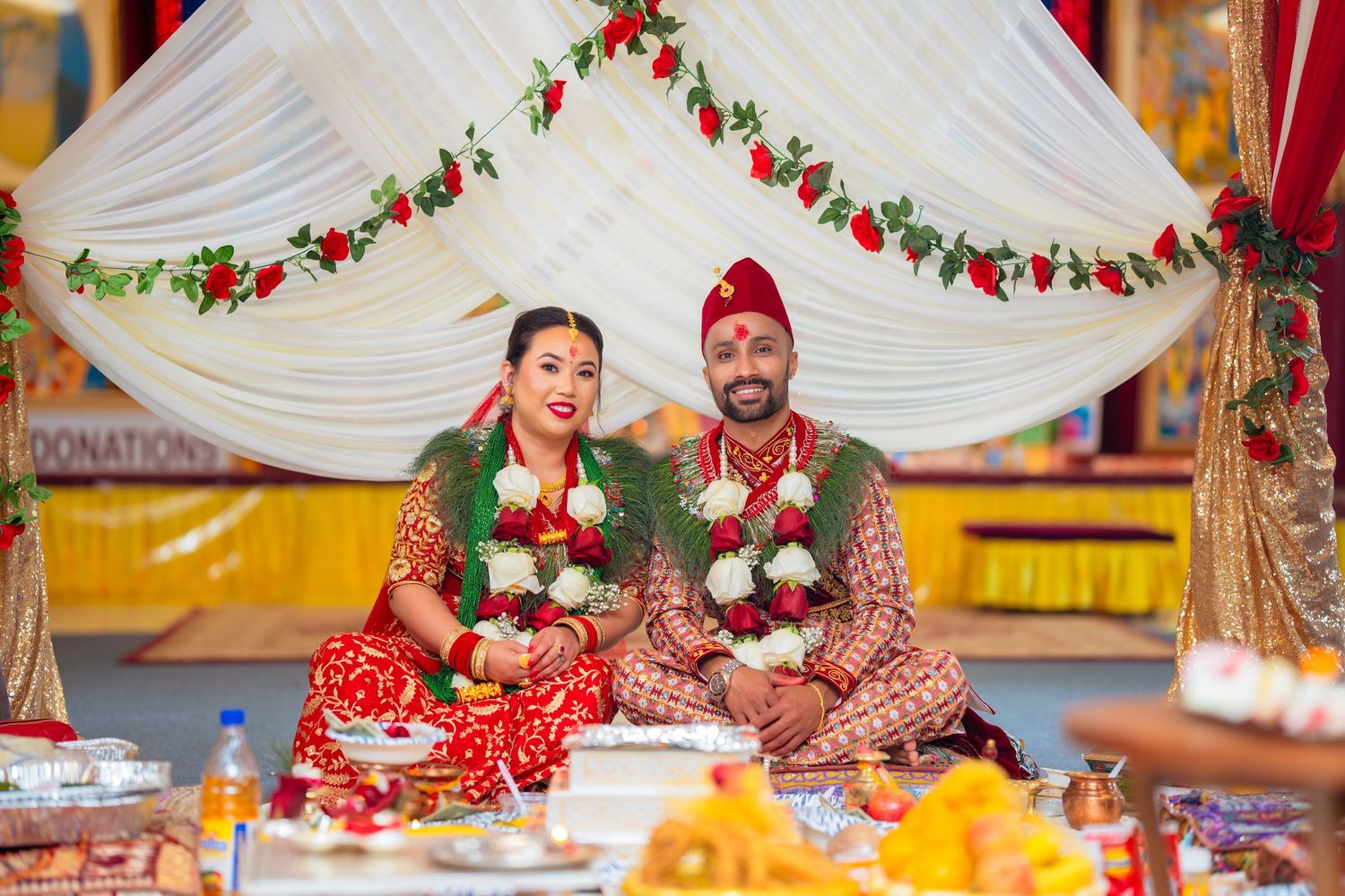 Newlyweds in traditional attire seated under floral mandap during Hindu wedding ceremony.