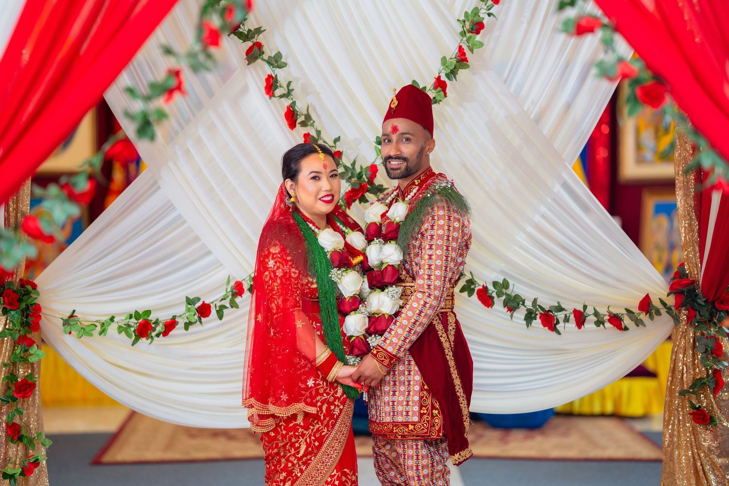 Traditional South Asian wedding ceremony with couple in red and gold outfits embracing under decorated mandap.