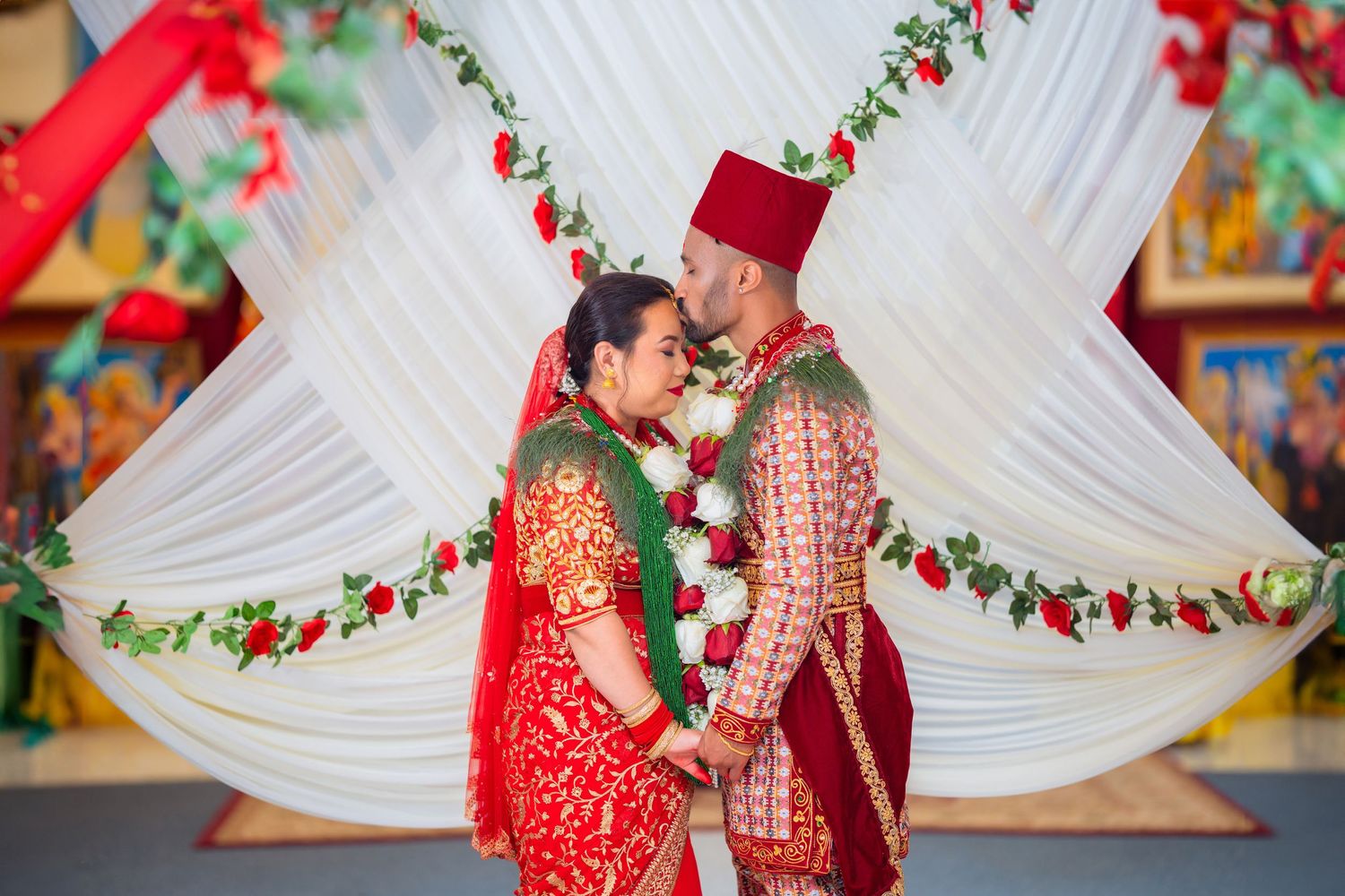 Couple in traditional red and gold wedding attire share an intimate moment under floral mandap decorations.
