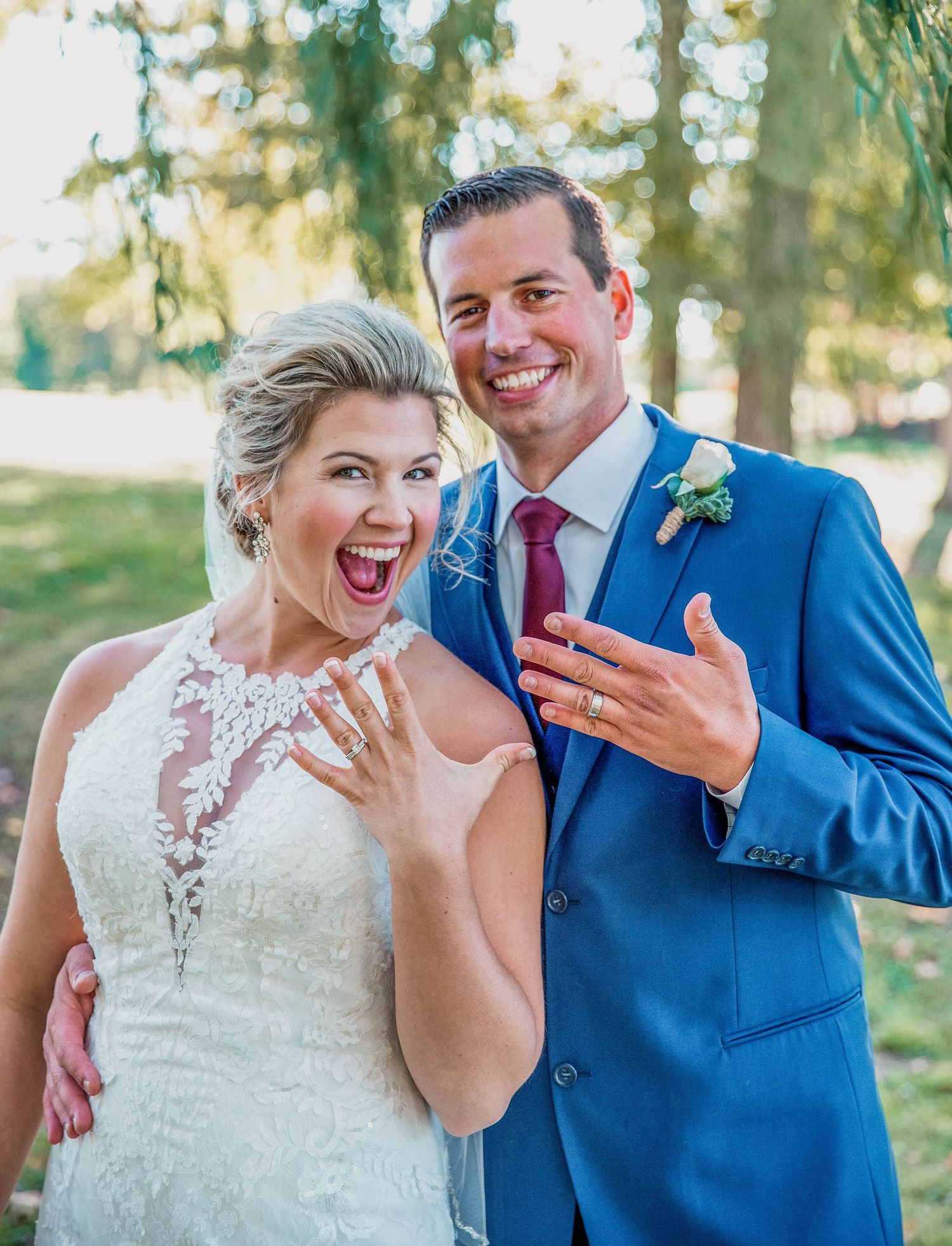 A newlywed couple shows off their rings with excited expressions in an outdoor setting.