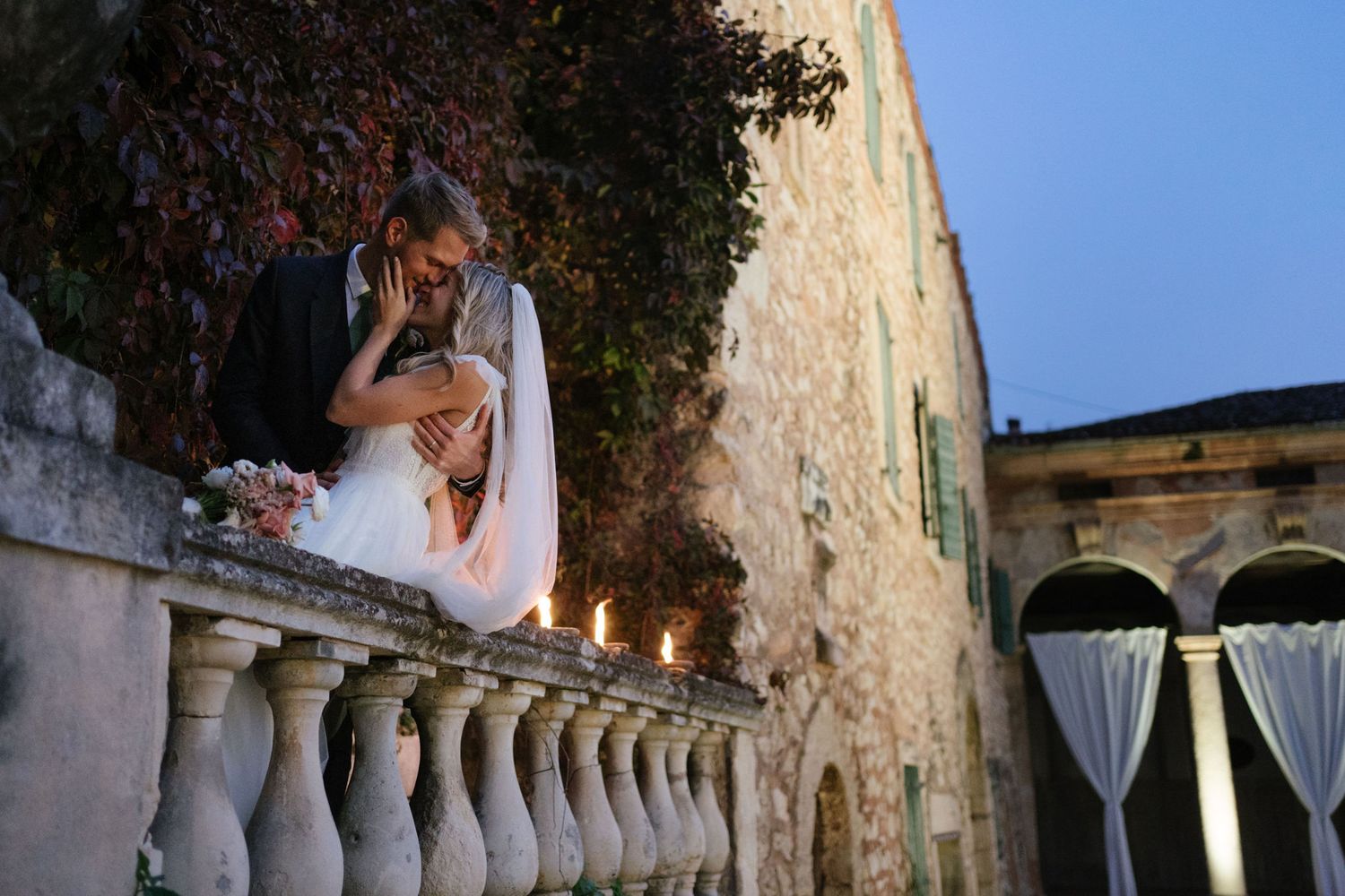 Romantic couple shares intimate moment on ornate balcony at twilight with candles glowing.