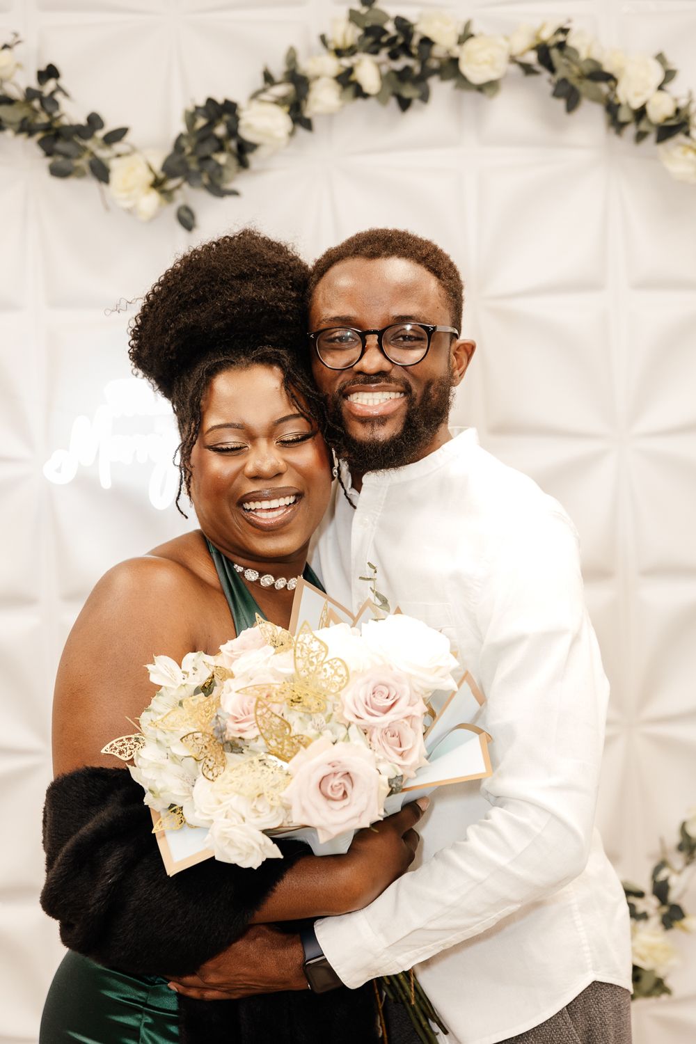A couple shares a joyful embrace while holding a bouquet of blush and cream colored flowers against a white backdrop.
