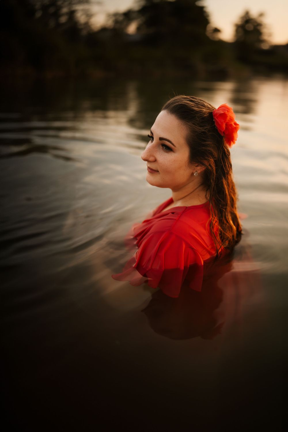 A woman in a red dress stands in dark water at sunset with a red flower in her hair.