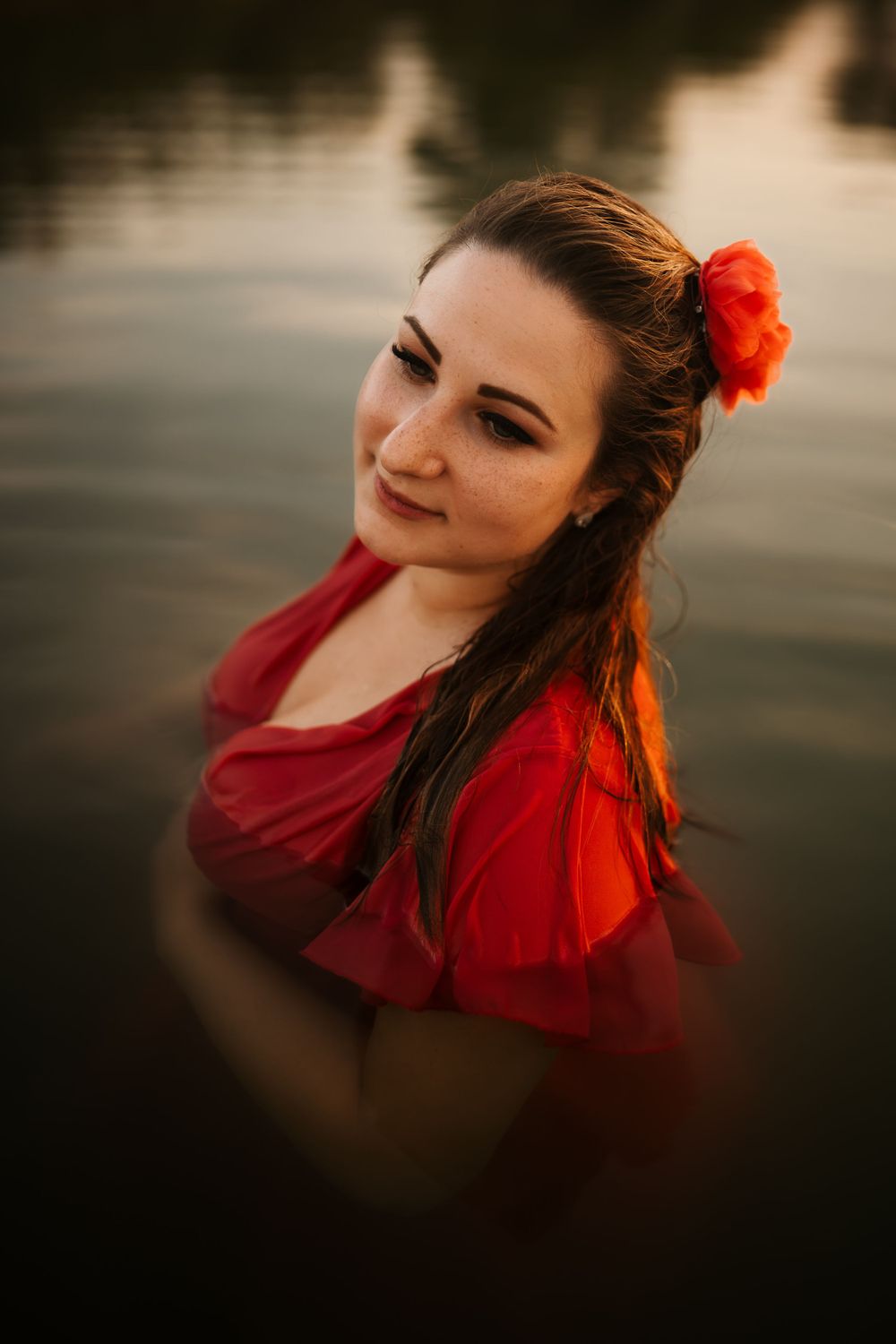 Woman in red dress with flower in hair stands in water during golden hour sunset.