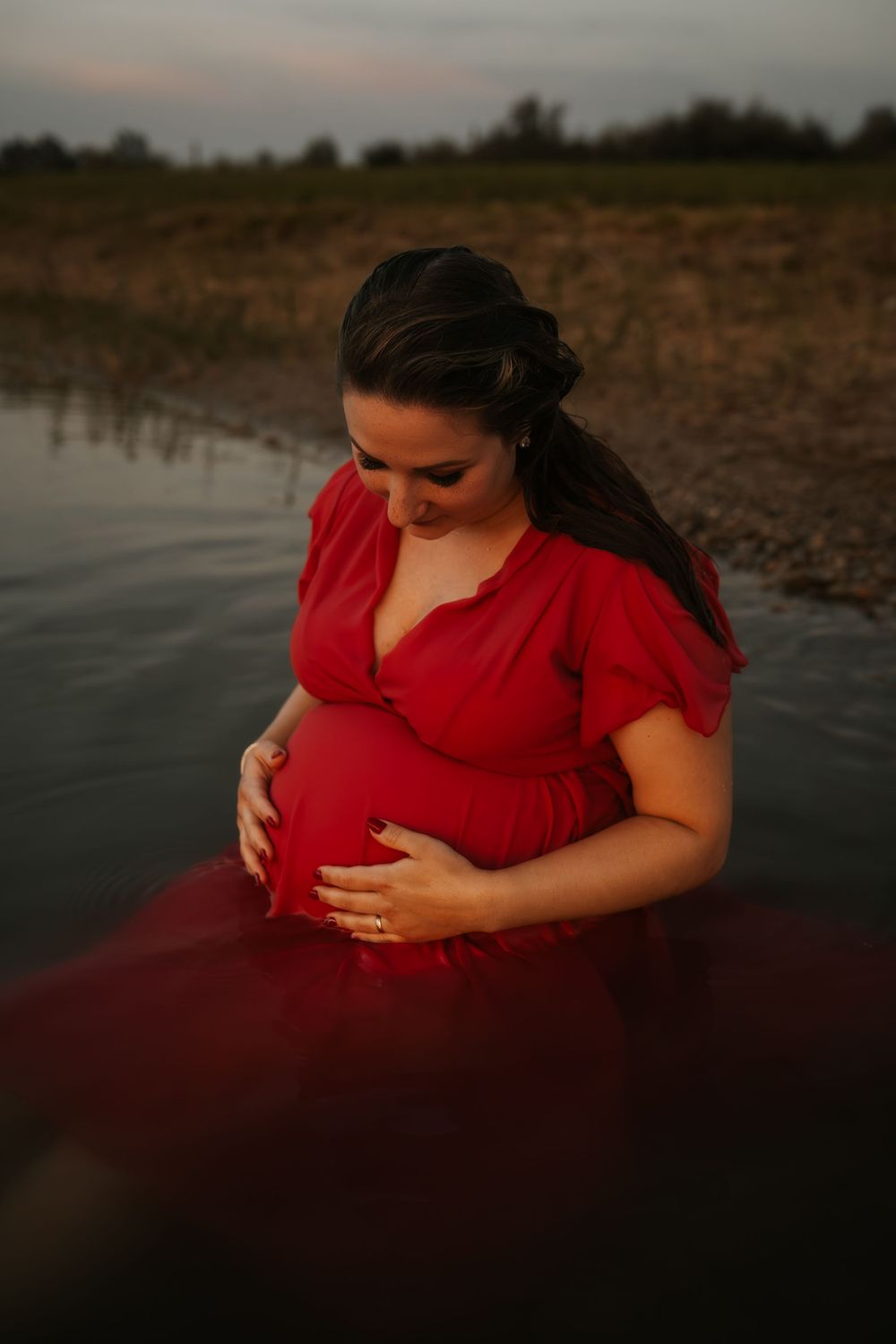 Expectant mother in red dress cradles baby bump while standing in water at dusk.