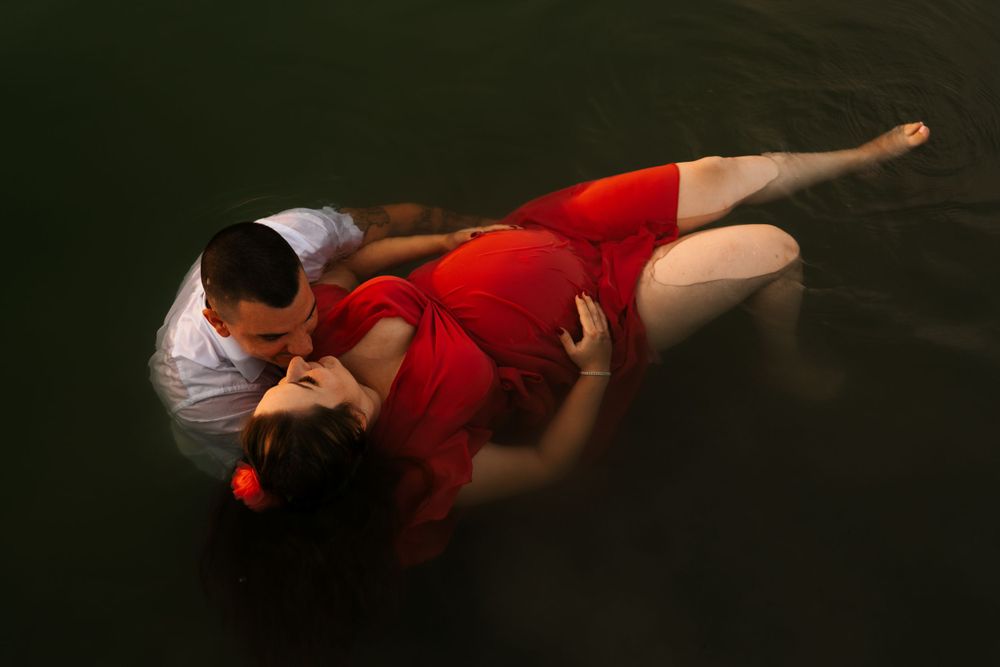 Couple in red dress and white shirt pose together in dark waters during dramatic photoshoot.
