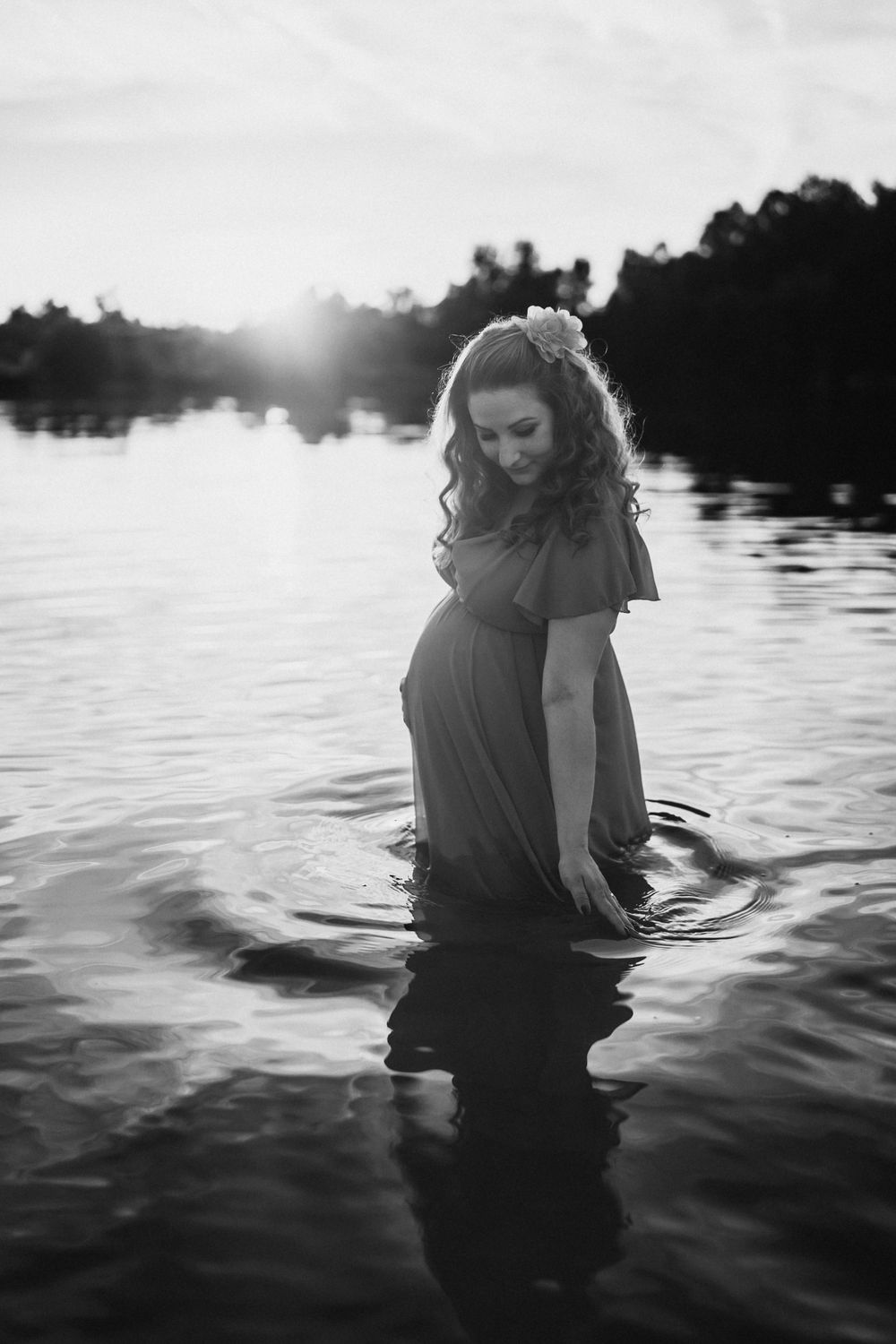 Black and white photo of pregnant woman standing in peaceful lake waters.