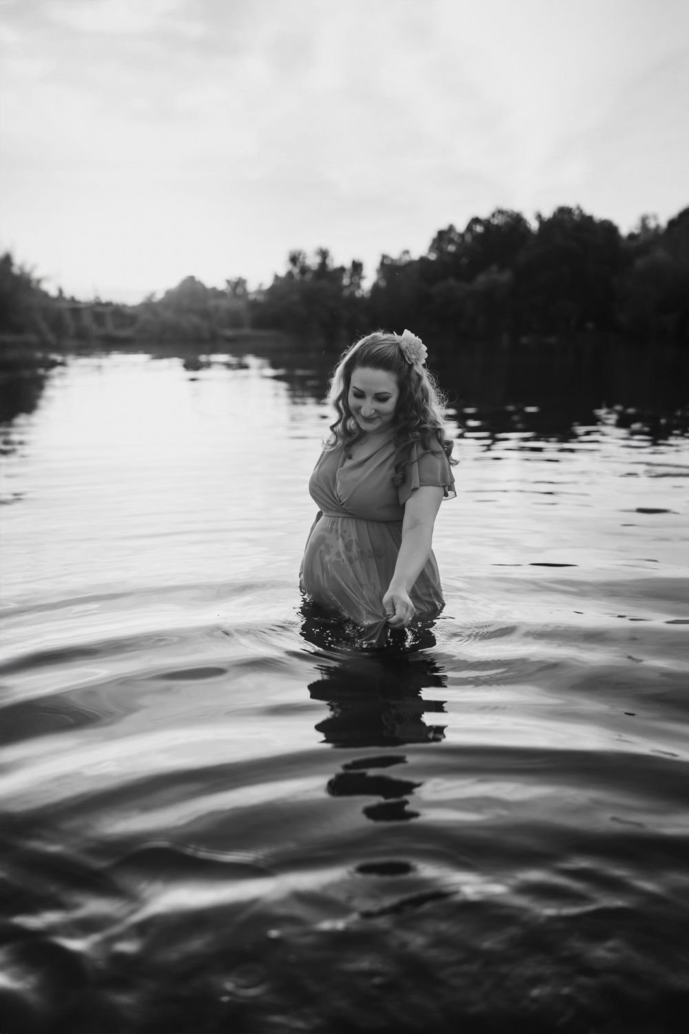 Joyful expectant mother stands in rippling water during black and white maternity shoot.
