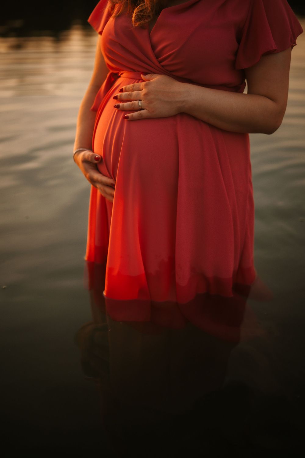 Close up of hands cradling pregnant belly in red dress by water at sunset.