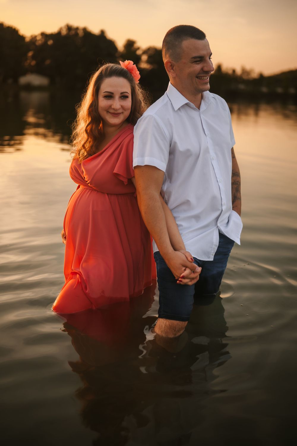 A couple holds hands while standing in water during sunset, wearing red dress and white shirt.