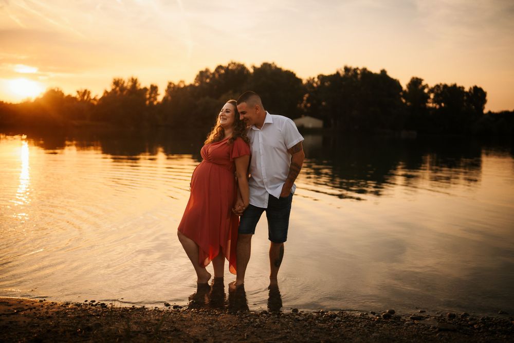 Happy couple poses together by lake during sunset maternity photoshoot.