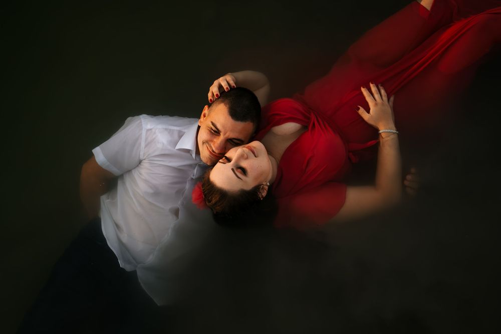 A couple in white and red clothing float together in dark water during a romantic photoshoot.