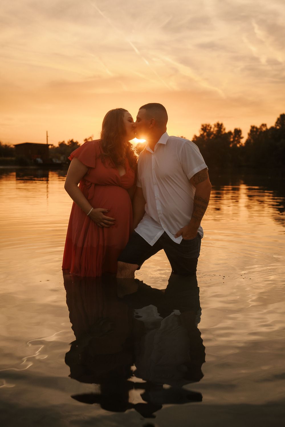 Silhouetted couple shares intimate moment in water during sunset maternity session.